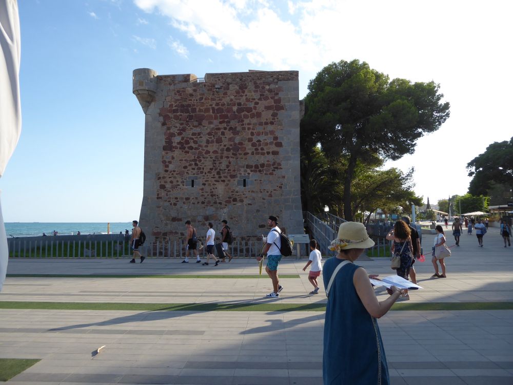 Una torre de vigilancia de la costa. En primer plano una persona consulta un plano. Mas atrás varias personas se dirigen hacia la playa. Cielo azul con nubes blancas