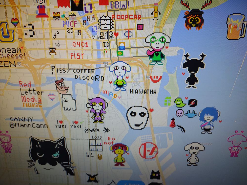 Photo of a monitor showing wplace and a map of Milwaukee filled with various pixel art, including several togore themed Deltarune characters and "I <3 yaoi/Yuri" sprite text