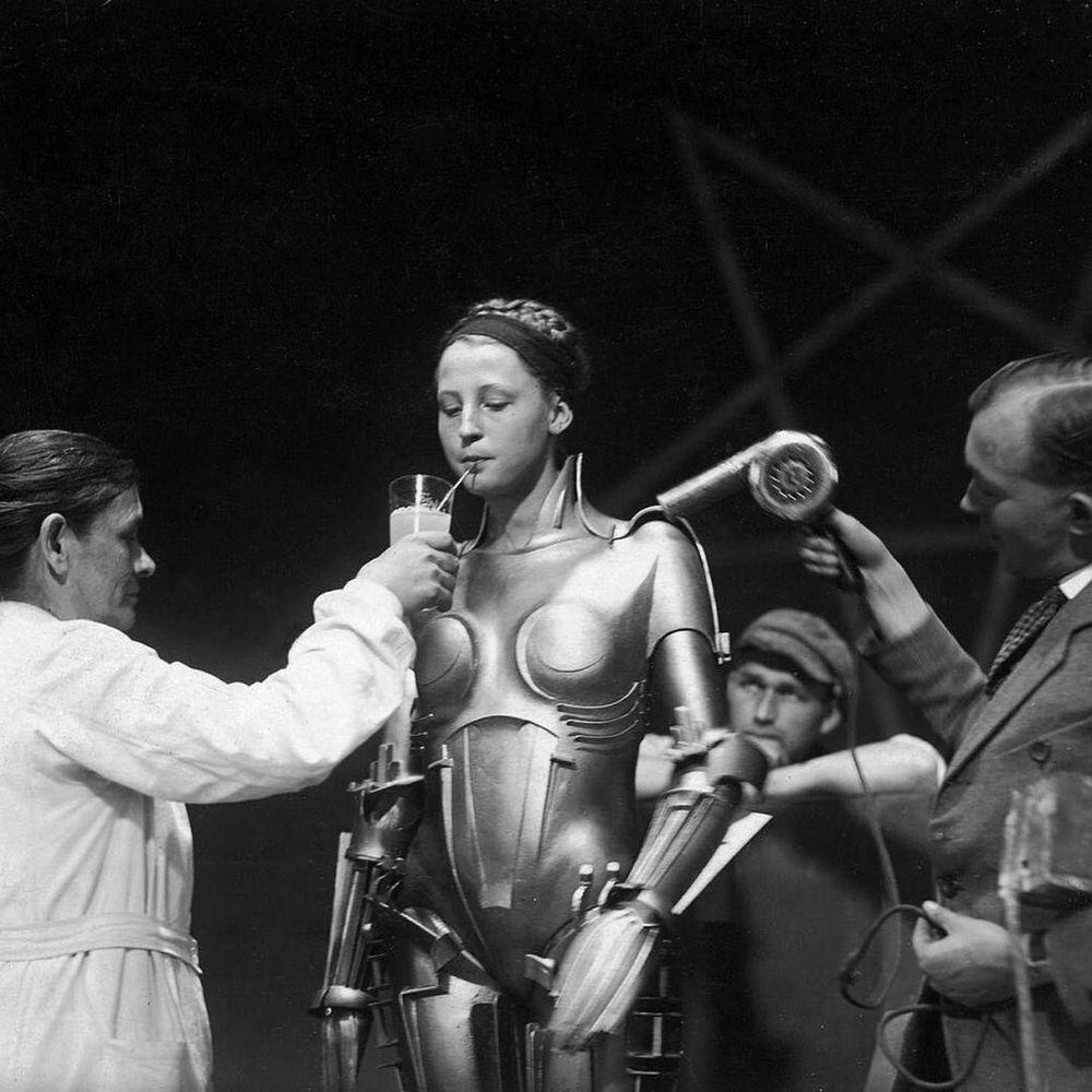 Brigitte Helm halfway in the Maria robot costume from Metropolis, being handed a cup of water or something to drink out of while someone else appears to cool her off with some kind of hairdryer
