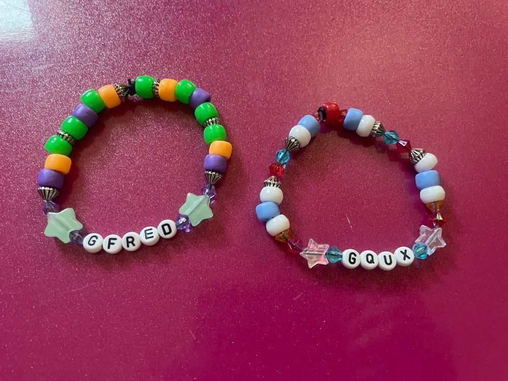 Two bead bracelets. One says GQUX and is white, red, yellow and blue, and the other says GFRED and is purple yellow and green.