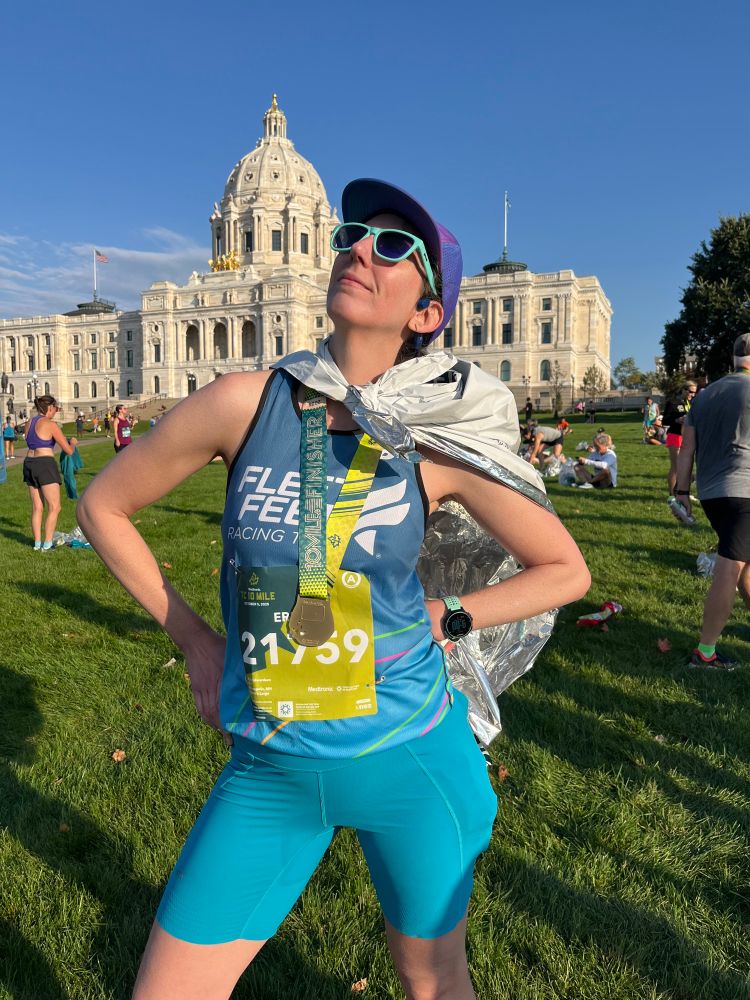 White woman in running gear including finisher’s medal and Mylar blanket blowing behind her, standing in front of Minnesota state capitol building