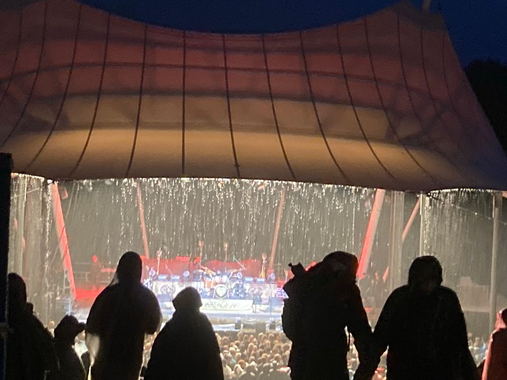 Eine überdachte Freilichtbühne, beleuchtet, von der das Wasser fließt und davor starker Regen 