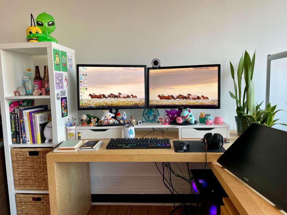 Photo of a tan wood L shaped desk in front of a white wall with two computer monitors showing a wallpaper with horses running in a field. To the left of the desk is a tall thin white bookshelf with multiple art books and decorative soda bottles. There are some small art prints on the side of the shelf and a green alien hugging a plush Perforce bee on top. On the desk to the right of the monitors are two snake plants. There is a large Huion tablet screen to the right on the L part of the desk. Under the monitors is a small white shelf with various Pokemon plushes and Kirby figures.