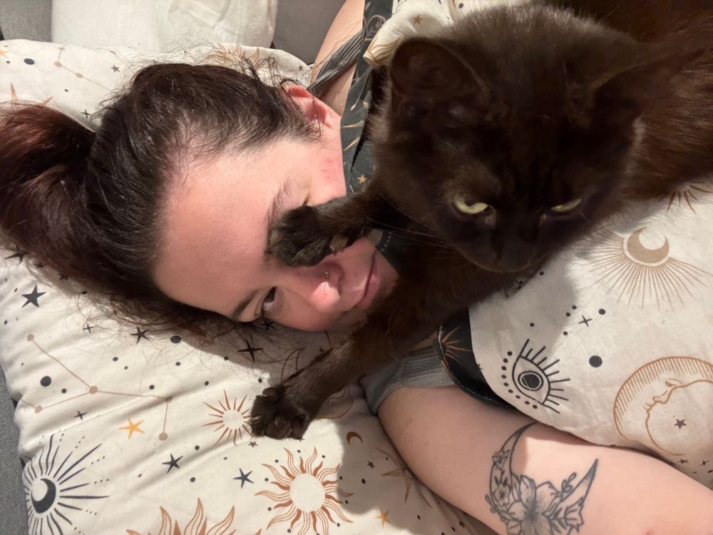 a brunette is laying in a bed with white celestial bed sheets. Their black/brown cat is laying on their chest, with her paw covering the persons eye