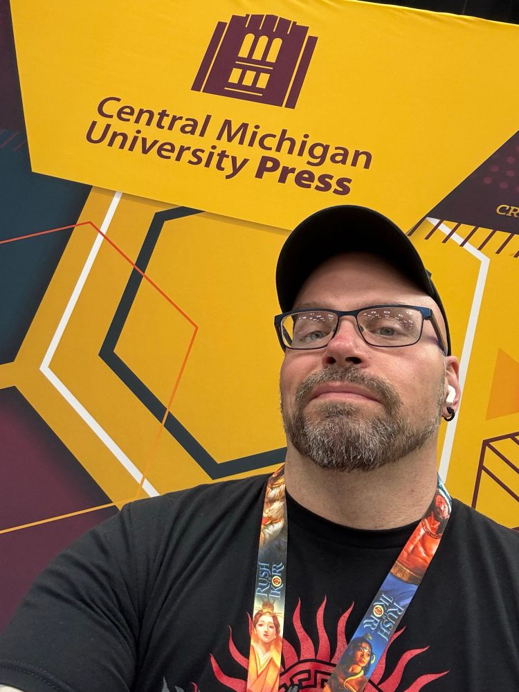 My semi-smiling face, in a selfie pose, with the CMich Press Gen Con banner behind me.