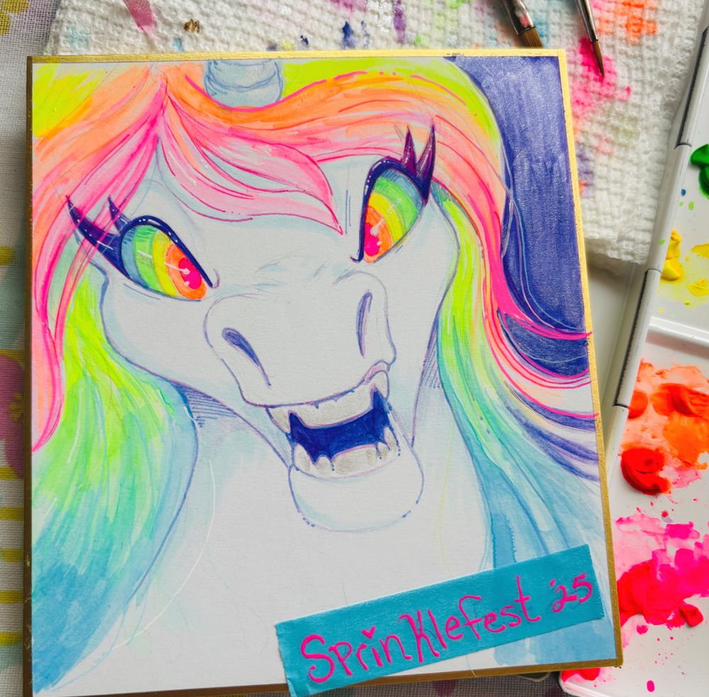 A photo of a watercolor sketch of a white and rainbow unicorn on a shikishi board with paintbrushes and a paint pallet slightly in frame. The unicorn has neon rainbow hair and eyes and is snarling angrily. The signature at the bottom on a square of blue tape says Sprinklefest 2025.
