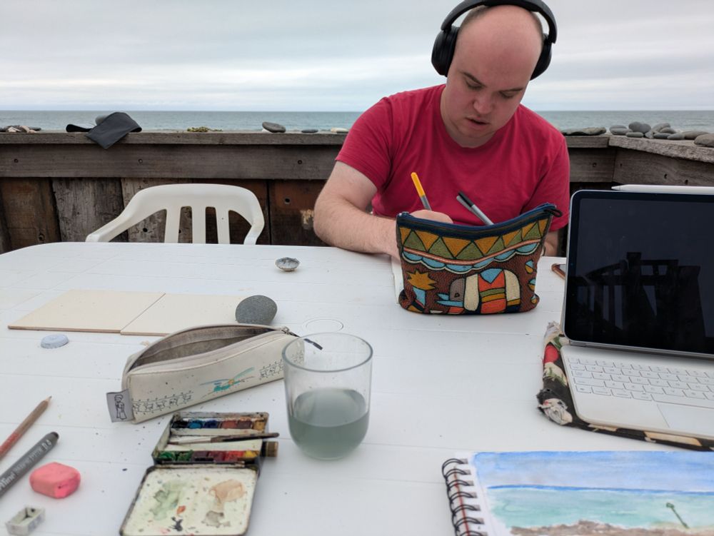 Photo of max with headphones drawing at plastic garden table. On the table : Max's pen bag, my iPad and watercolours. In the background : the Irish Sea. 