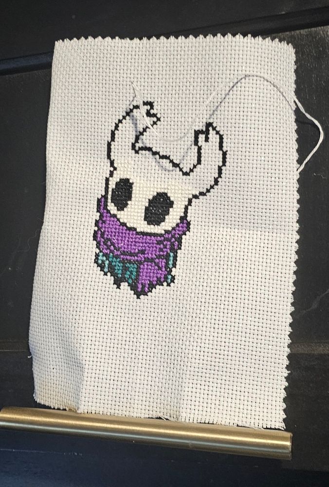 Counted cross stitch of a hollow knight video game character with a purple scarf on with some blue underneath. 