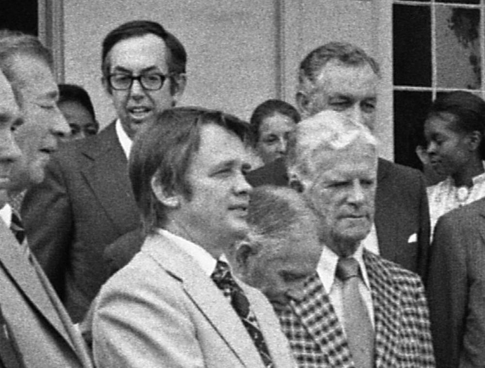A close up of a photo on the porch at Arlington House on August 5, 1975, showing part of a woman's face behind two men, with only her eyes and a portion of her dress visible