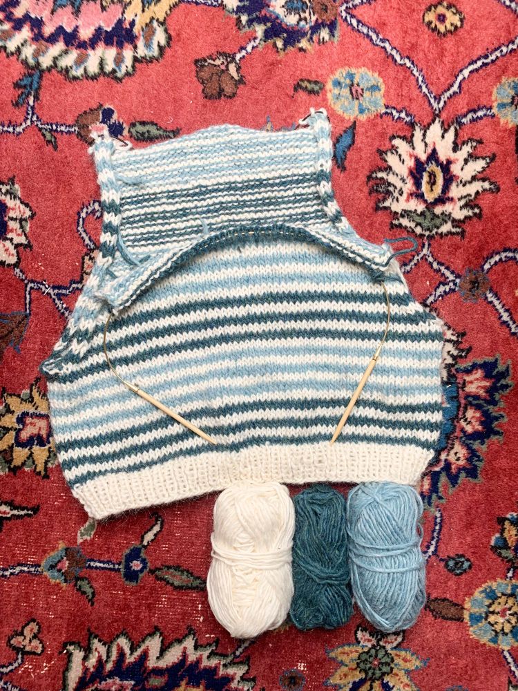 A half-finished hand knit striped slipover lying on the floor on a colorful rug.  