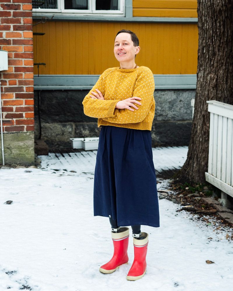 Kaisa is standing outside in the snow wearing a yellow knitted sweater with white dots, blue skirt and red rubber boots. Kaisa is a double amputee wearing prosthetics on both legs. 