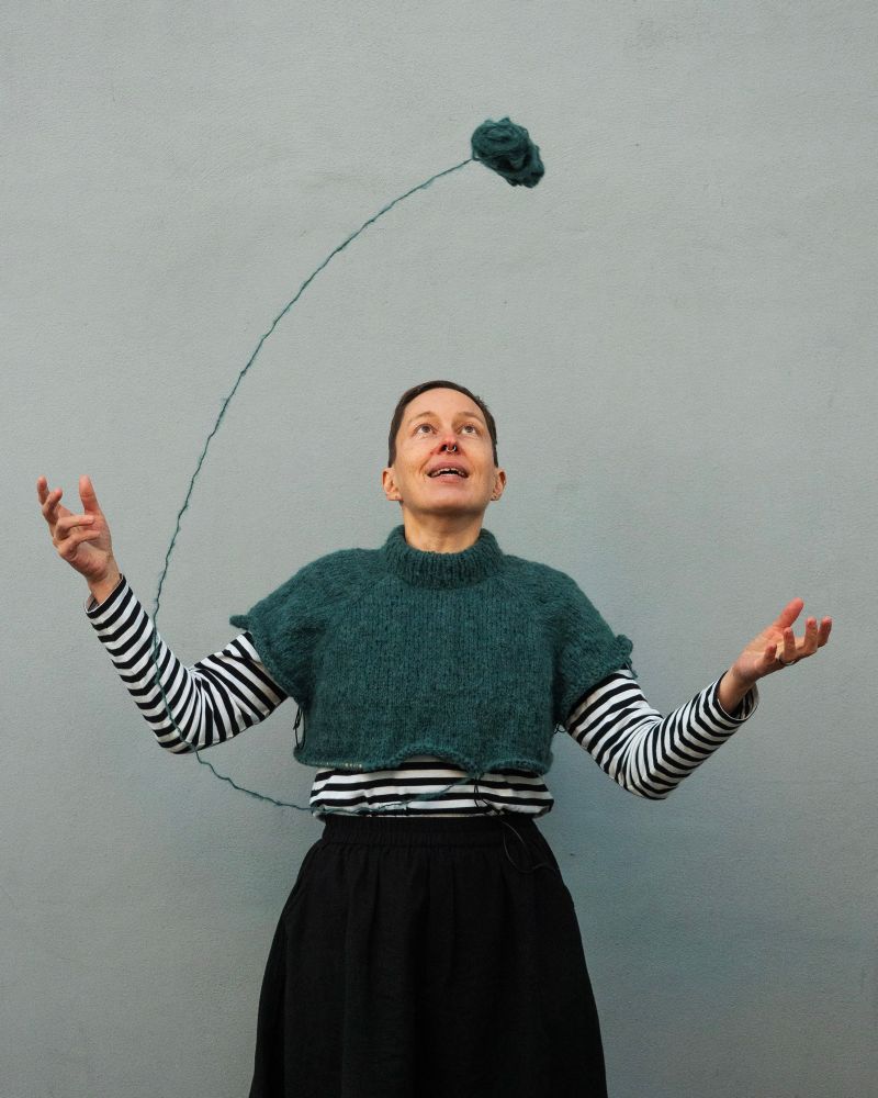 Kaisa is wearing a half-finished hand knit sweater and throwing a skein of yarn from one hand to the other. 