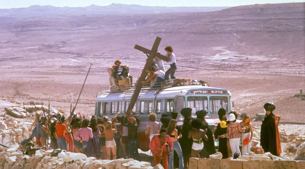 A group of young people hoist a large wooden cross onto a bus, which is parked in a vast desert.
