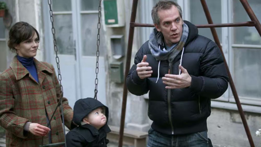 On a Parisian street, a woman and a small boy look on as a man in a puffy coat explains something to someone off-camera, gesturing with his hands. 