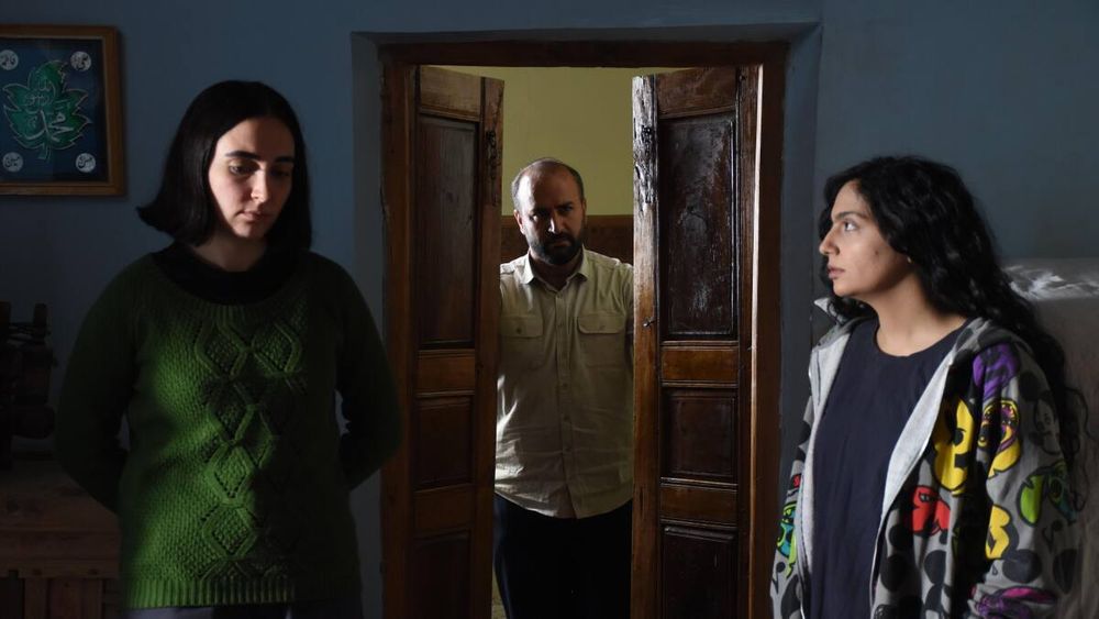 Two young women stand uncomfortably on either side of a doorway as an older man closes the doors on them.
