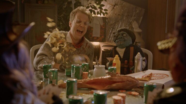A blond man with glasses reacts as a handful of potato chips fly off the kitchen table in front of him. There's a weird little puppet friend sitting in the chair to his left.
