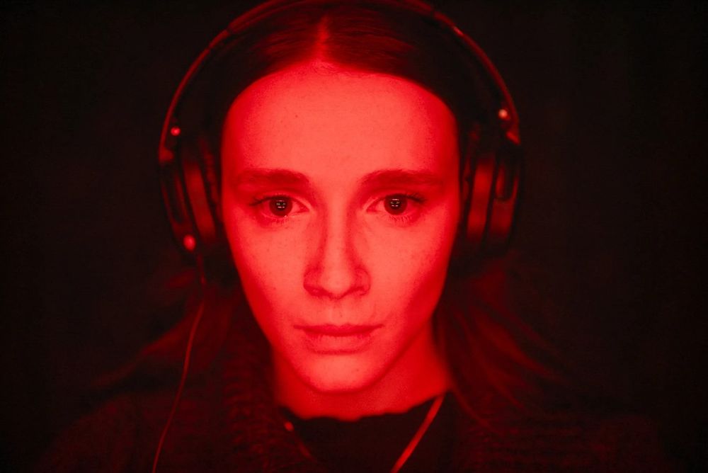 Bathed in orange-red light, a young woman wearing headphones stares intently at something just below the camera. (Juliette Gariepy in a still from RED ROOMS.)