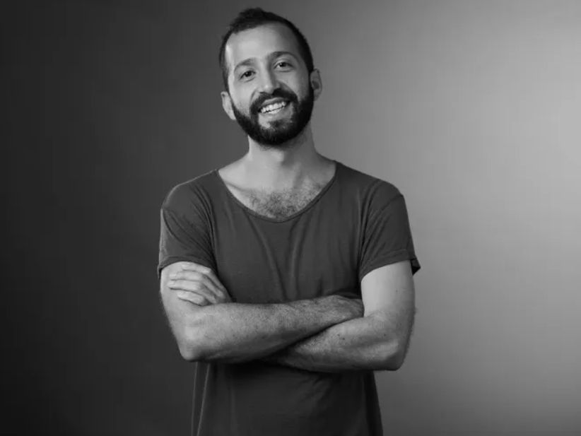 A bearded man in a t-shirt smiles for the camera, his arms crossed. The image is in black and white.