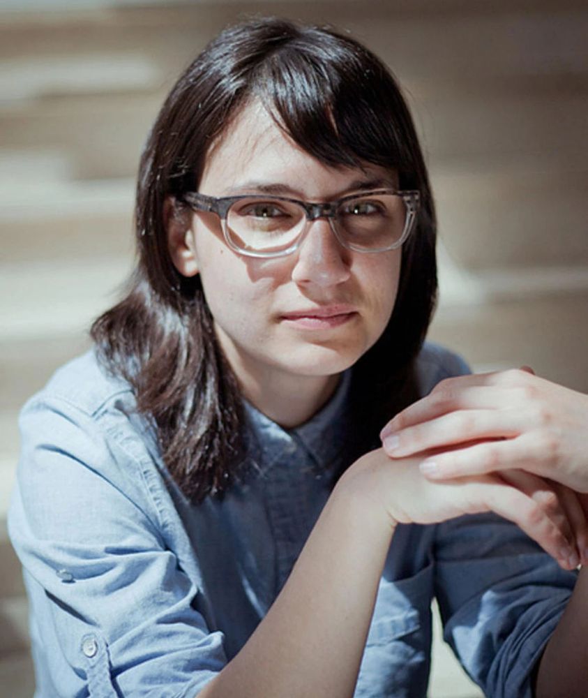 A young woman with dark hair and glasses looks at the camera, wearing a blue Oxford shirt with the sleeves rolled up.
