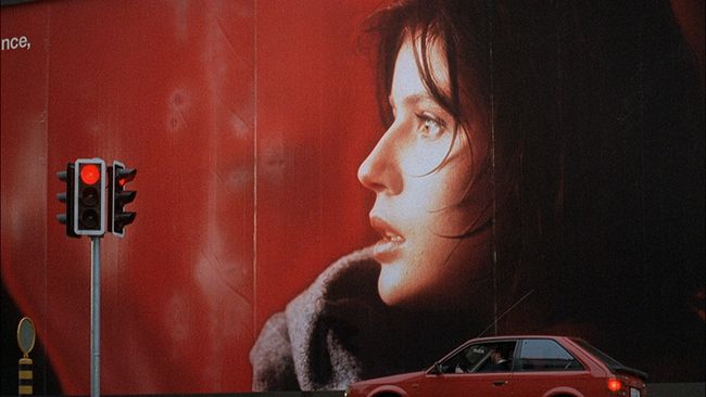 A small red car sits at a red light at a street corner. The building in the background sports a massive image of a woman's face in profile, framed against an indistinct red background.

(Image is from THREE COLORS: RED.)