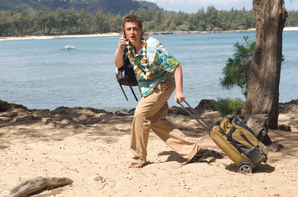 A man in a tropical shirt and khakis drags a rolling case across a sand beach, looking exactly like the famous silhouette image of Bigfoot. If Bigfoot had luggage.