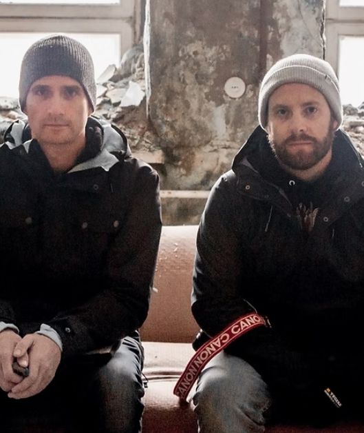 Two men -- one with a little stubble, the other with a beard -- sit on a couch in a decrepit building. They both wear toques and parkas. 