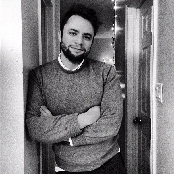A bearded man leans against a wall in a hallway, his arms crossed in a comfortable pose.