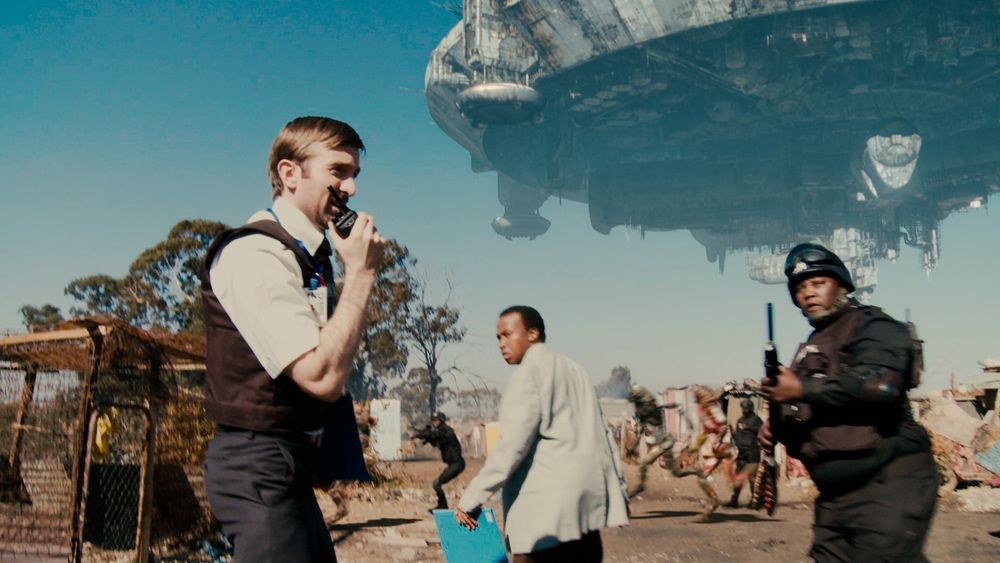 A man speaks into a walkie-talkie  as two passersby -- one in civilian clothes, another in paramilitary gear -- glance at him. A massive saucer-shaped ship is visible in the sky behind them. 

(Image is from Neill Blomkamp's DISTRICT 9.)