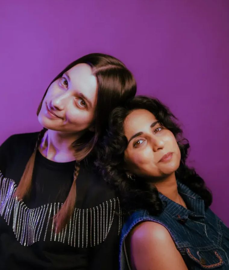 Two young women lean against each other against a purple backdrop in a promotional photo. They look happy!