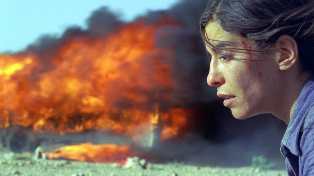 A shocked woman stares down at the ground as something burns in the distance behind her. (Image is a still from Denis Villeneuve's INCENDIES.)