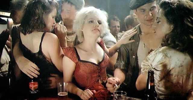 At a 1950s-era bar, a platinum-blonde woman stares into the middle distance while couples smoke and drink on either side of her.