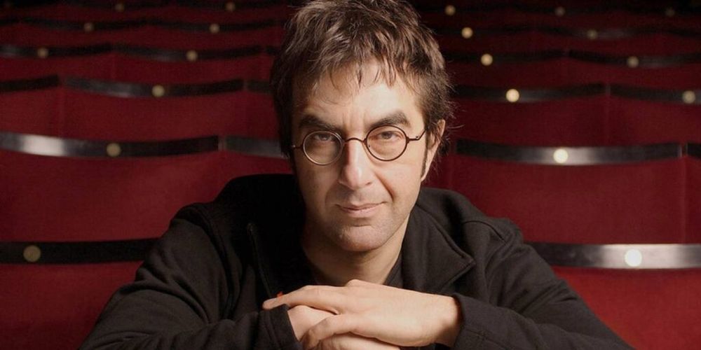 Sitting in a theater, a man wearing a black shirt leans forward and looks intently into the camera.