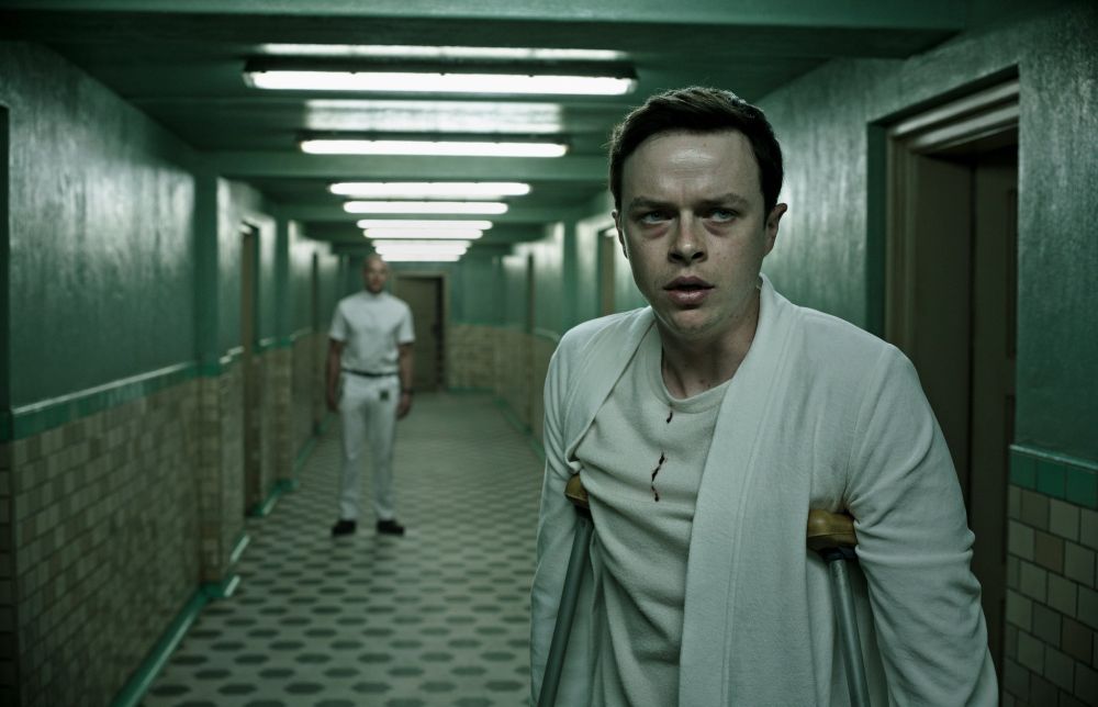Supporting himself with crutches, a man stands in a vaguely institutional hallway looking nervous and confused. He's dressed in a white robe over a white T-shirt, which has a trail of blood running down the center. An orderly stands in the distance behind him, also dressed in white, arms at his sides. The image has a sickly green tint to it.