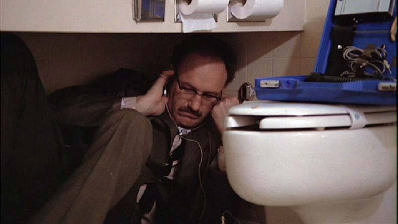 A balding man in a brown suit crouches uncomfortably underneath a sink in a hotel bathroom, listening intently to whatever's playing in his headphones. A freshly sanitized toilet is directly to his left.