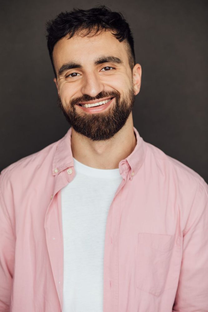A bearded man smiles for the camera. He's wearing a rose-colored shirt over a plain white T.
