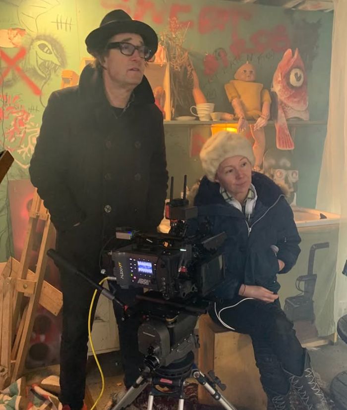 A man and woman perch behind a video camera in a messy space. 