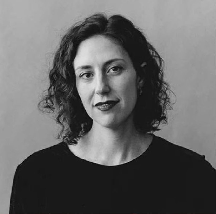 A black-and-white image of a woman facing the camera. She has shoulder-length dark hair and wears what looks like a black T-shirt.