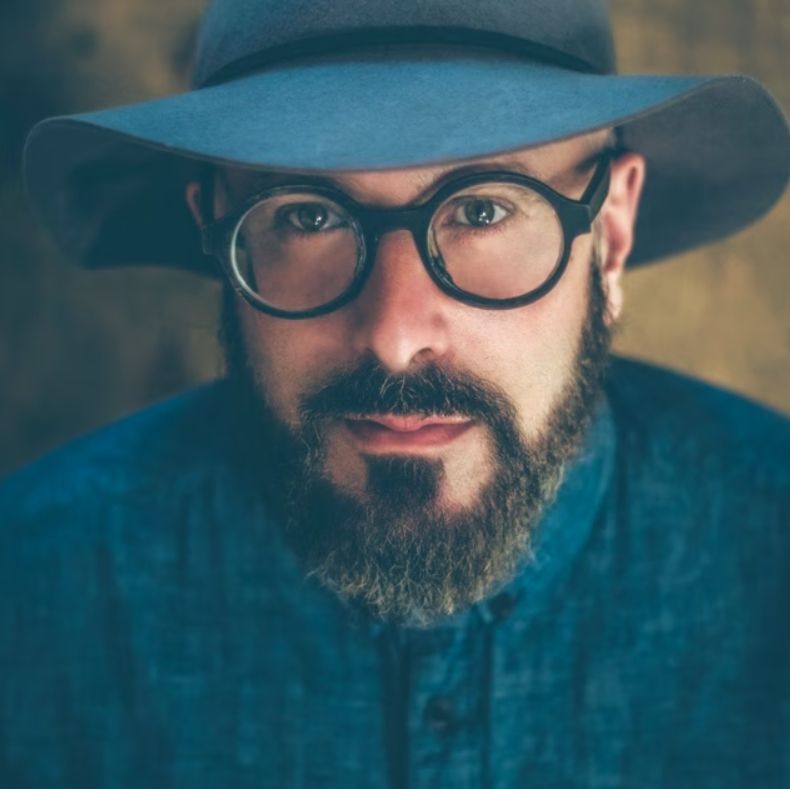 Wearing large round eyeglasses, a blue hat and a blue shirt, a bearded man stares straight into the camera. 
