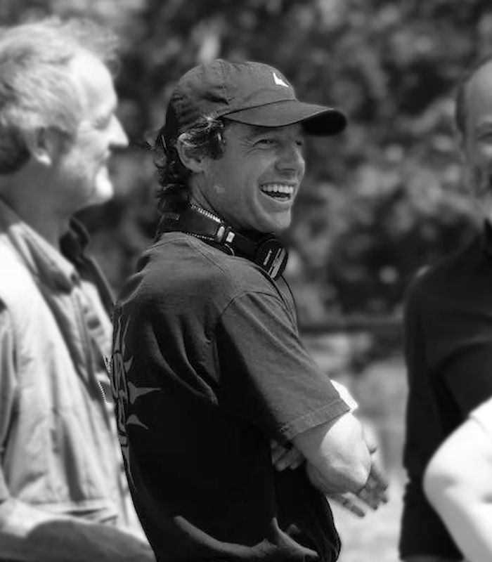 A black-and-white photo of a young man in a ball cap and T-shirt, with headphones around his neck, smiling broadly at something off-camera.