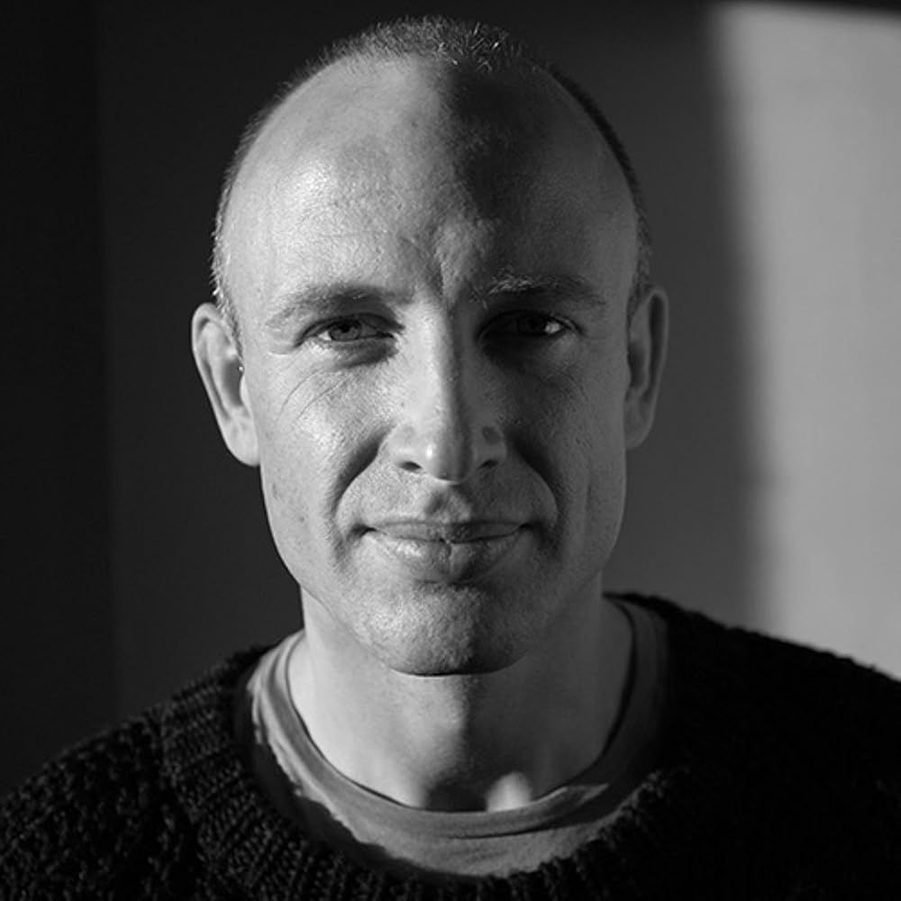 A man looks directly at the camera. He has close-cropped hair and wears a sweater over a T-shirt.