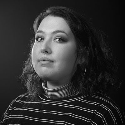 Wearing a black sweater with white stripes over a black-and-white striped turtleneck, a woman looks at the camera in three-quarter profile. The image is in black and white, too.