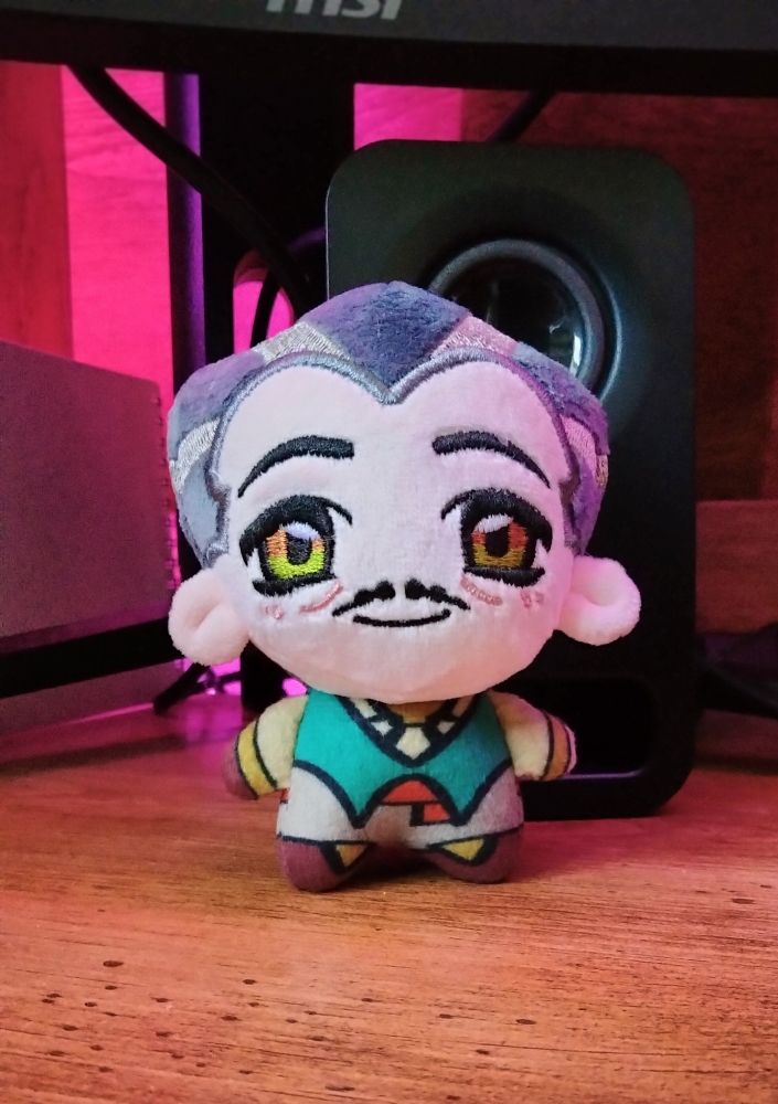 A plush keychain of Emmrich from Dragon Age, sitting on a desk. He is very polite and supportive of you.
