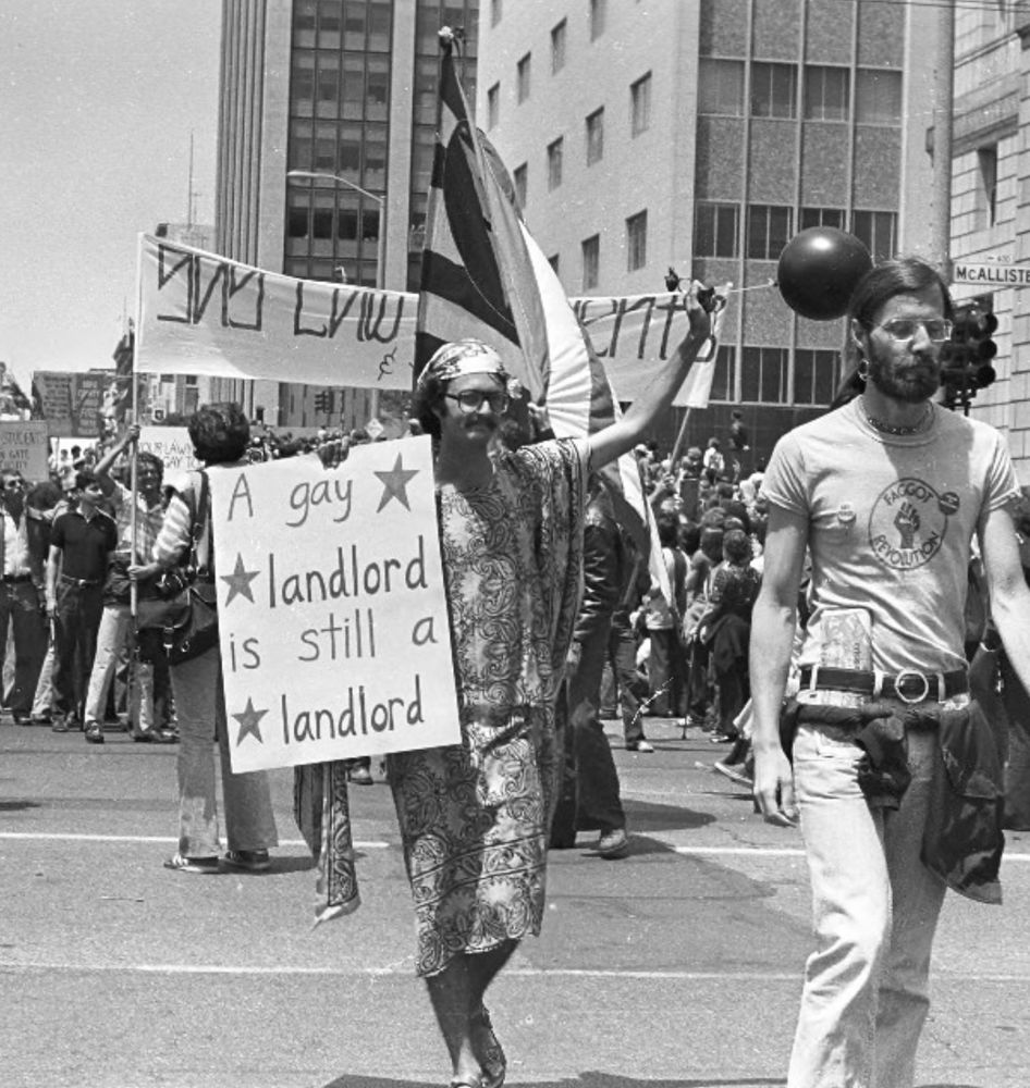 Man in a parade holding a sign that says “A gay landlord is still a landlord.” Another man in front of him is wearing a “faggot revolution” t-shirt. 