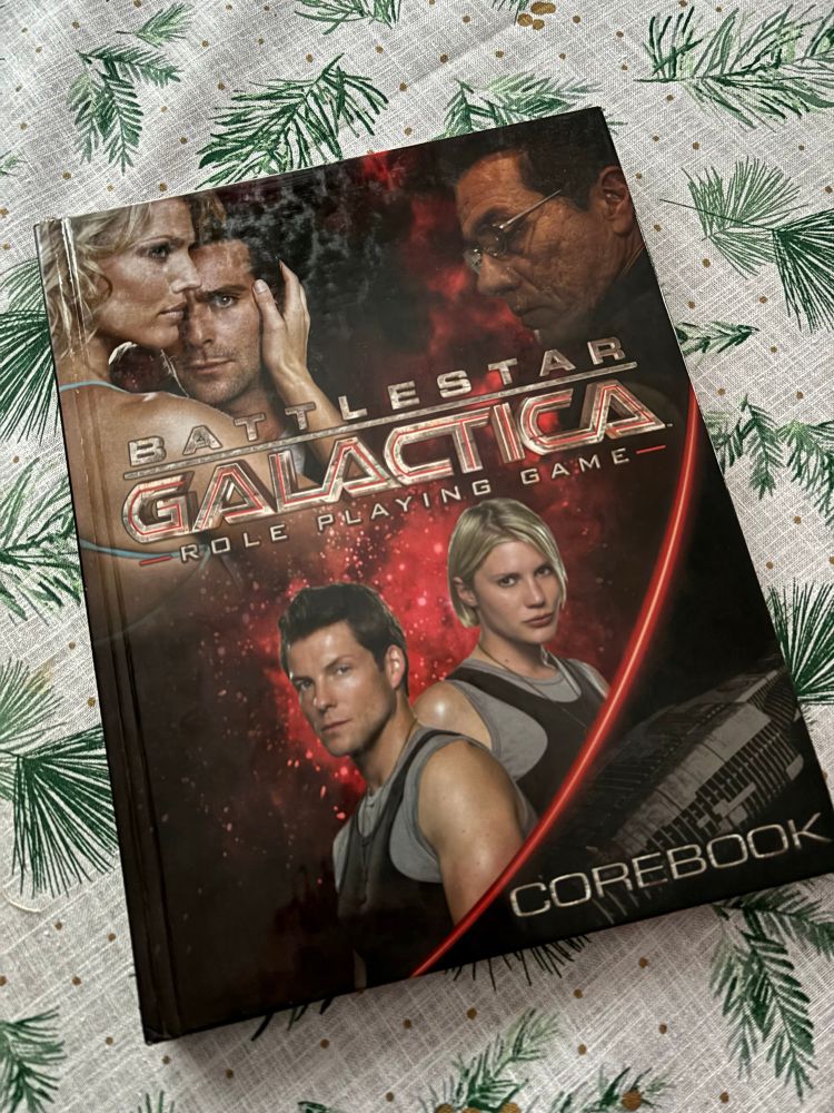 A photo of the Battlestar Galacatica Role Playing Game laying flat on a table with a white and green winter’s tablecloth. 