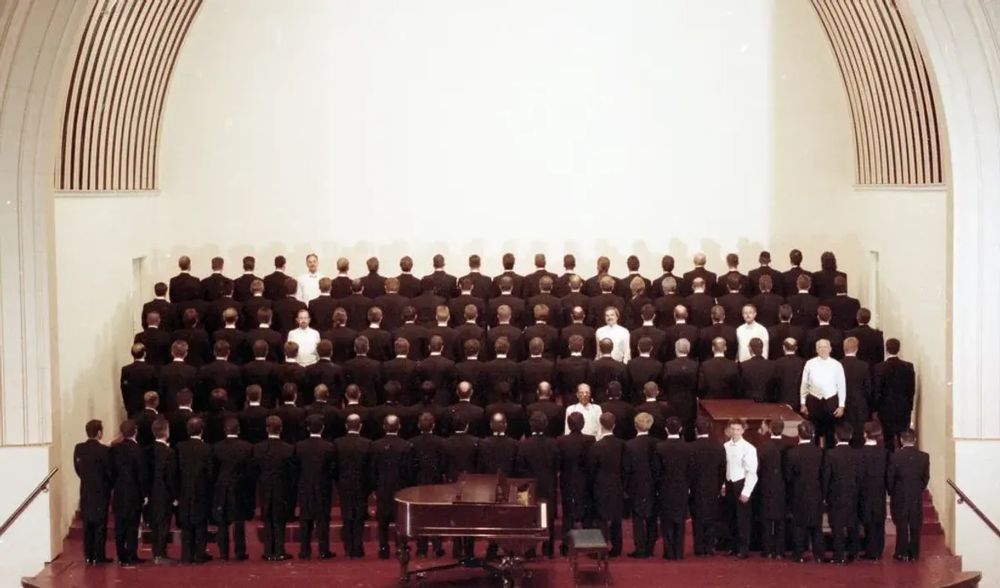 A portrait of the San Francisco Gay Men's Choir in 1993. The seven surviving members of the original choir face the camera, dressed in white; they are all seemingly young or middle-aged. The other 115 members face backwards, dressed in black, representing all of the original choir members who had died of AIDS as of 1993.