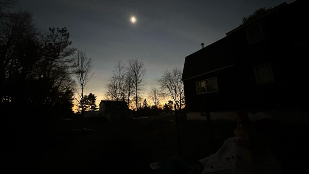 Total solar eclipse wide shot of a dark back yard and dark house in front of a completely eclipsed sun with sunset colors on the horizon.