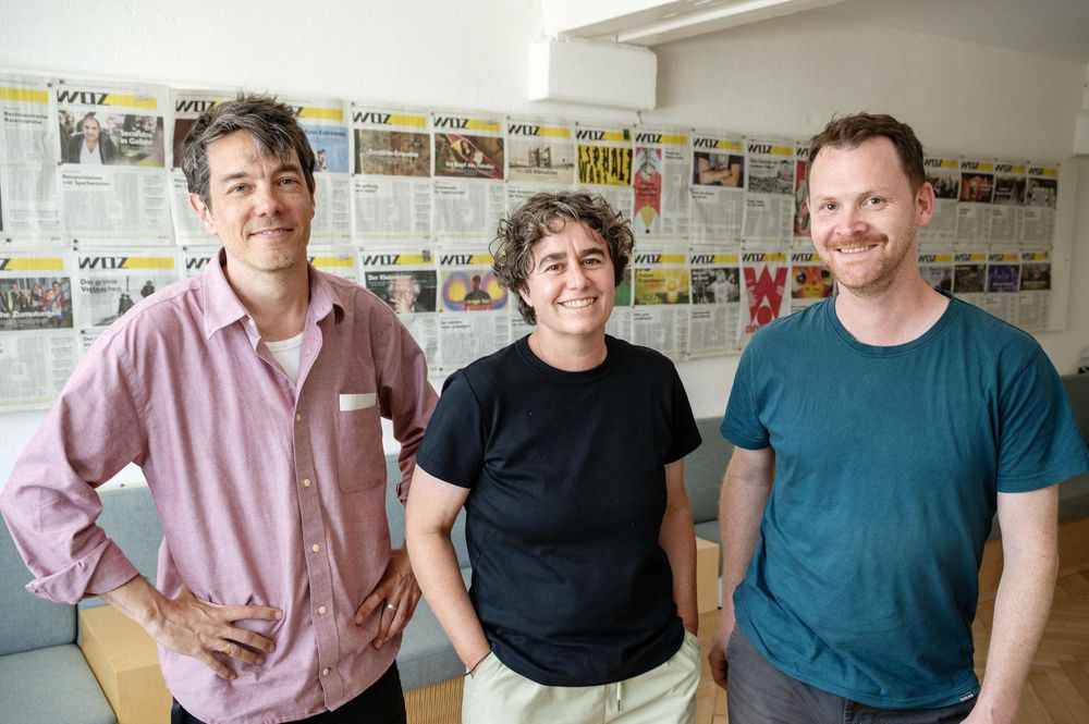 Florian Keller, Daniela Janser und Renato Beck stehen lächelnd vor einer Wand, an der Titelseiten der WOZ hängen.