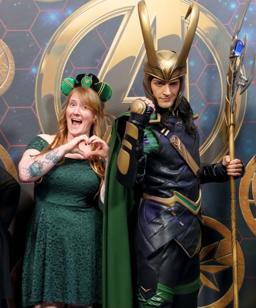 Zu sehen sind 2 Personen, links eine Frau im grünen Kleid die mit den Händen ein Herz formt, rechts ein Disney-Cast Member verkleidet als Loki