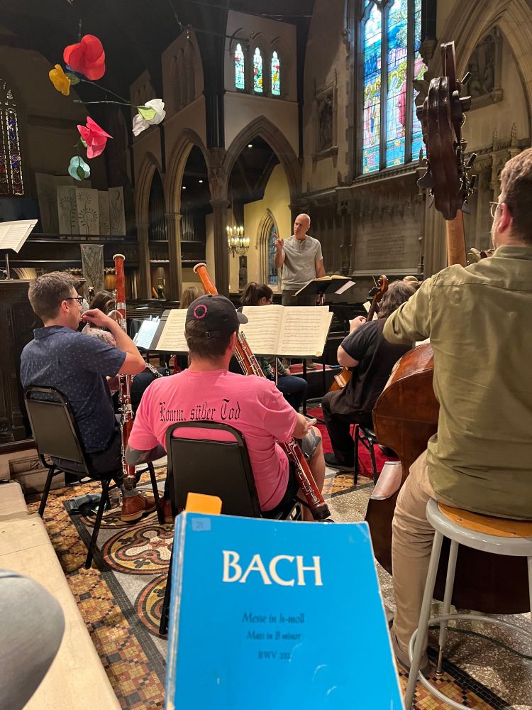 Picture of Emmanuel Music rehearsal; score of B minor Mass in foreground and conductor Ryan Turner on podium speaking to musicians.