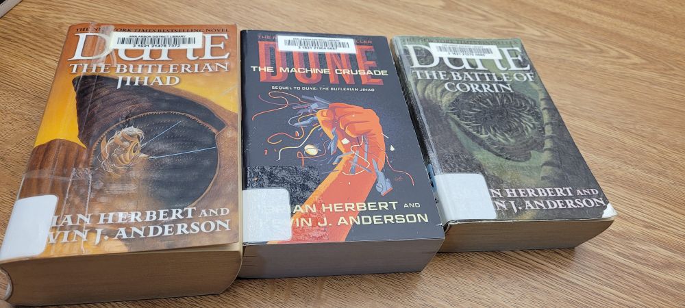 A picture of three library books next to each other on a faux wood table: Dune: The Butlerian Jihad, Dune: The Machine Crusade, and Dune: The Battle of Corrin
